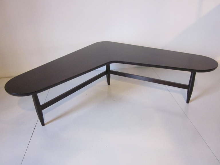 A  Flying V or L shaped wooden coffee table refinished in a dark and rich ebony satin ,sitting on three legs with cross stretchers.
