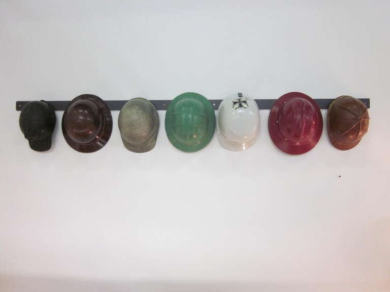A collection of seven vintage fiberglass working mens helmets included are Coal miner, Oil rigger, Racing car driver, Electrical Line worker, Soap Box Derby racer, Construction worker and Jockey. All hang on a custom made steel beam with build in