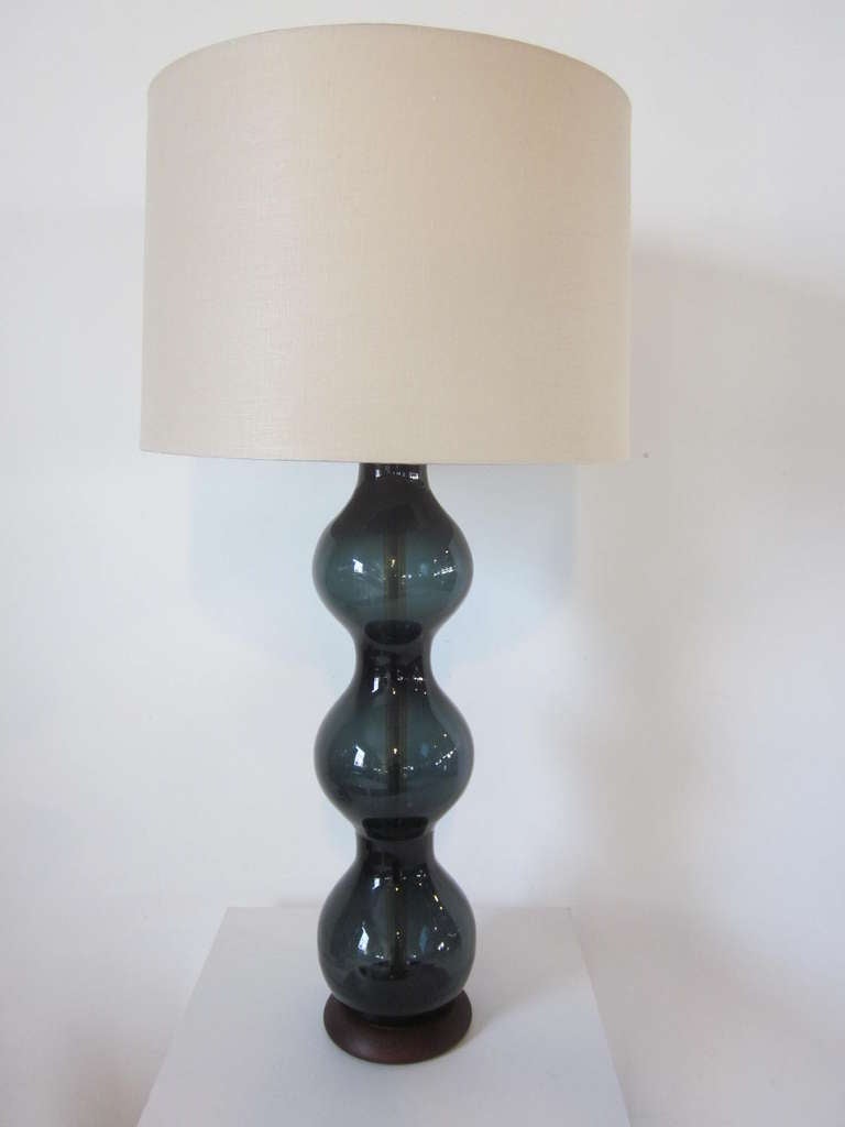 A artist blown Blenko glass table lamp in a steel blue tone sitting on a walnut wood base and topped by a linen shade.