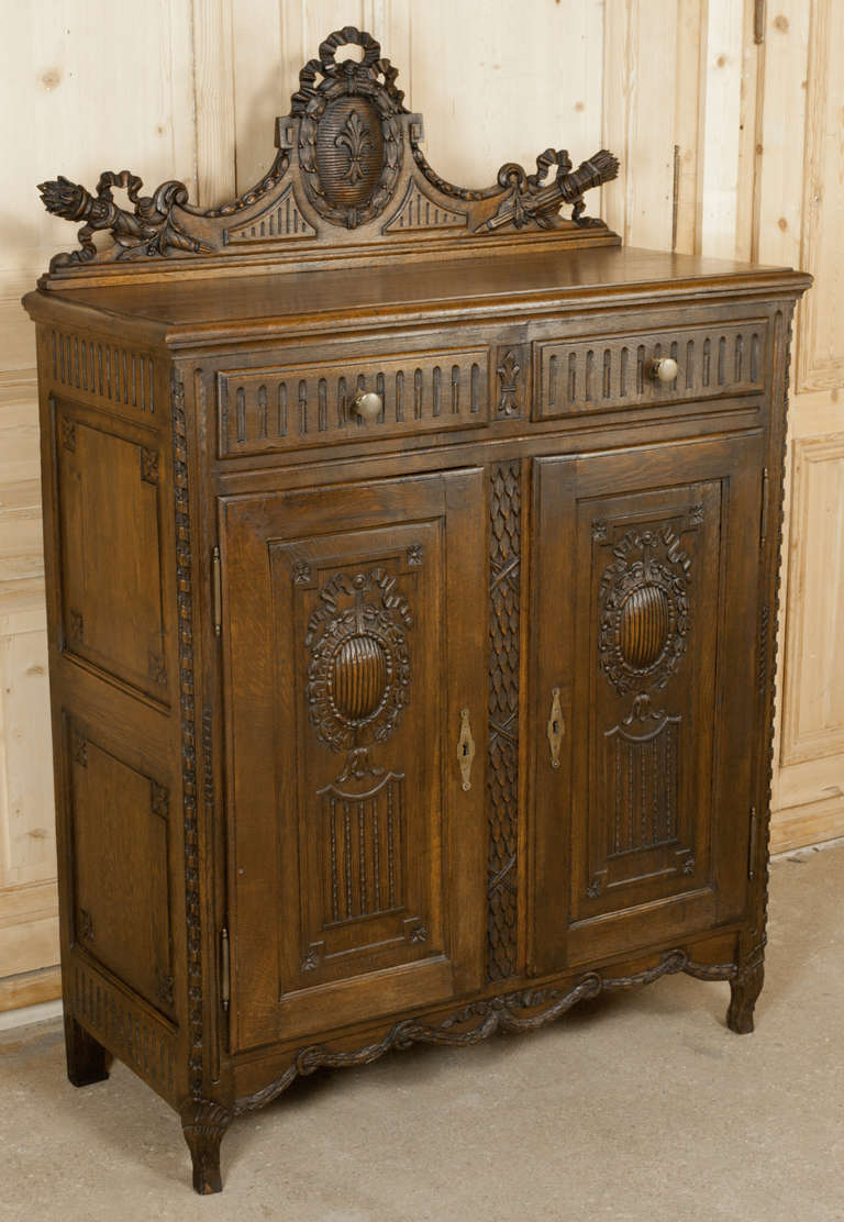 This Antique Rustic French Louis XVI Cabinet dates to the Louis XVI period, and features elaborate hand-carved detail including flowing ribbon, laurel wreaths, laurel swags, a fasci down the center stile and a backsplash carved with the royal fleur