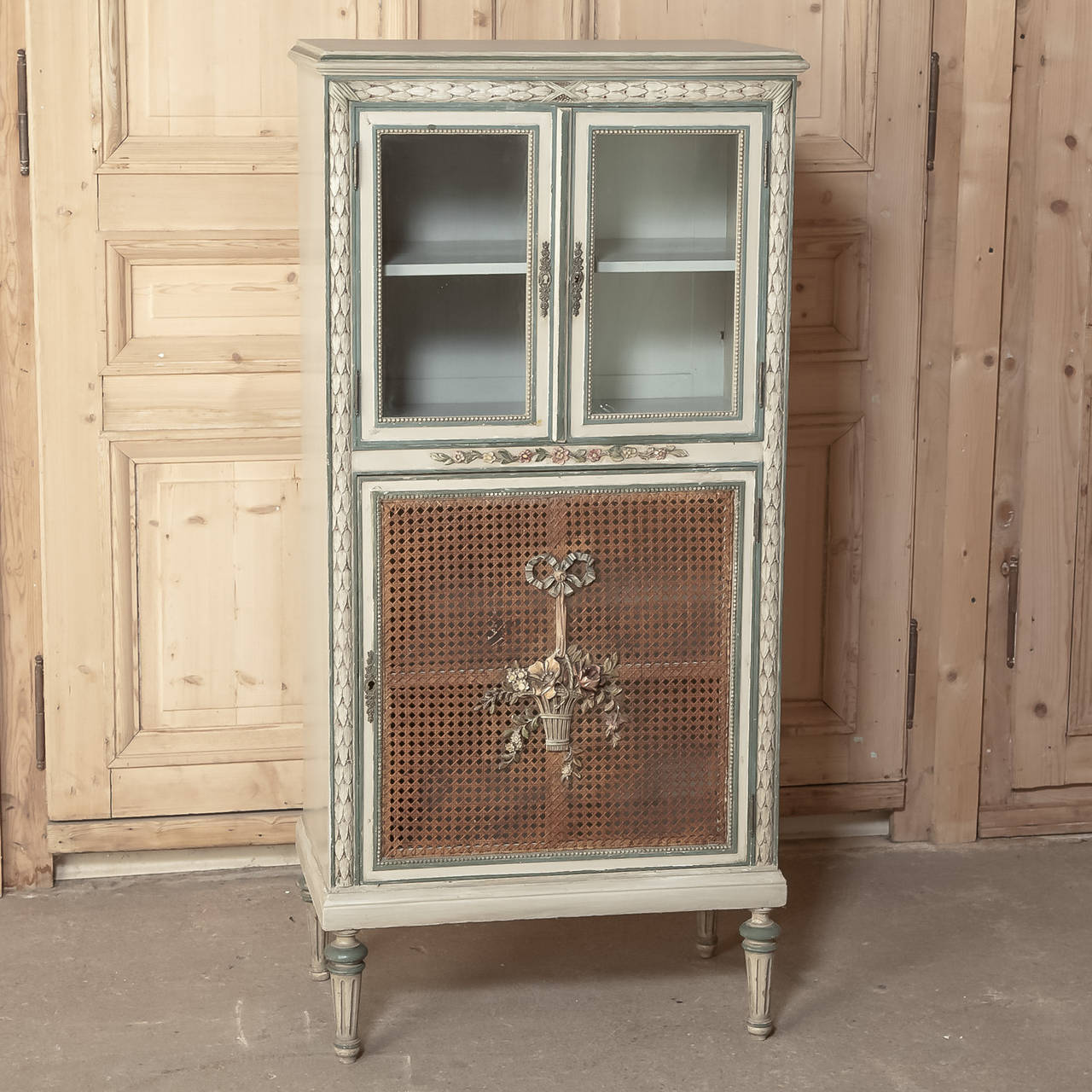 Boasting Classic styling, hand-carved embellishment, caned panel and display, all with original patinated painted finish, this charming little antique French Louis XVI vitrine allows one to display on top and store on bottom with style! Of