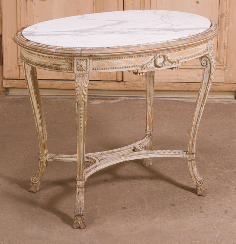 Boasting elaborate and fine detail in the carved embellishments that adorn the entire oval apron, gracefully scrolled legs and even the intertwined stretchers, this elegant Antique French Louis XVI Painted Marble Top Table has a wonderful distressed