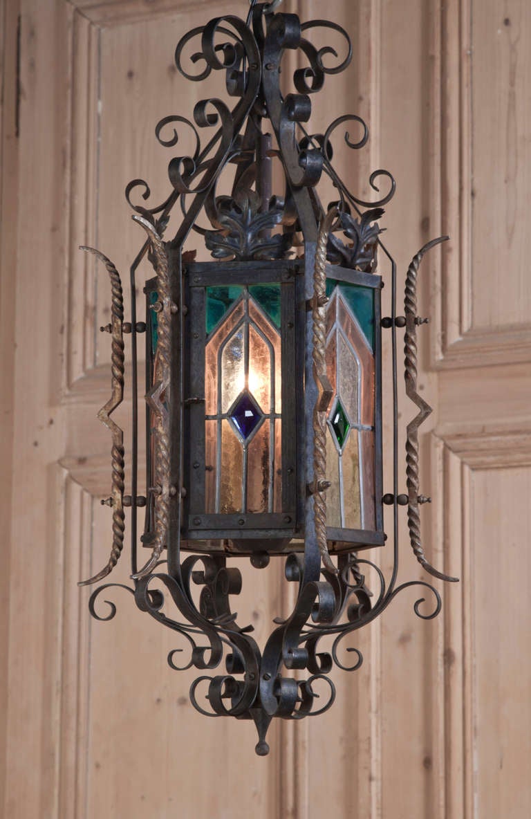 Wrought from iron into an intricately detailed Gothic showpiece, this lantern style chandelier has been fitted with stained glass panes with twisted and tapered rods on the corners highlighted in gold to create an intriguing source of light