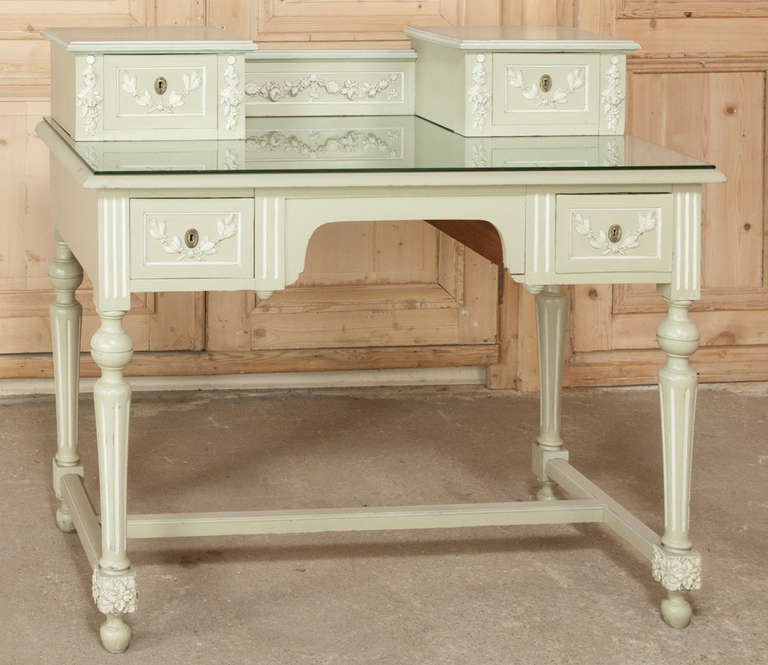 Perfect as a desk or a vanity, this little jewel features carved detail in the Louis XVI manner with two-toned patinaed paint, plus a glass top to protect the main writing surface. 
Circa 1900-1910. 
Measures 37H x 42W x 20.5D; surface 31H.