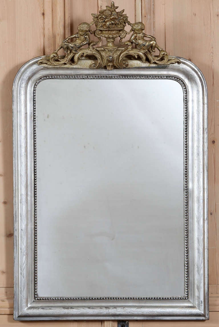 Featuring a classically understated Louis Philippe style frame with subtle embossing showing through the silvered finish, this mirror boasts an elaborate aged, golden finished cartouche on top depicting a pair of cherubs atop foliate flourishes