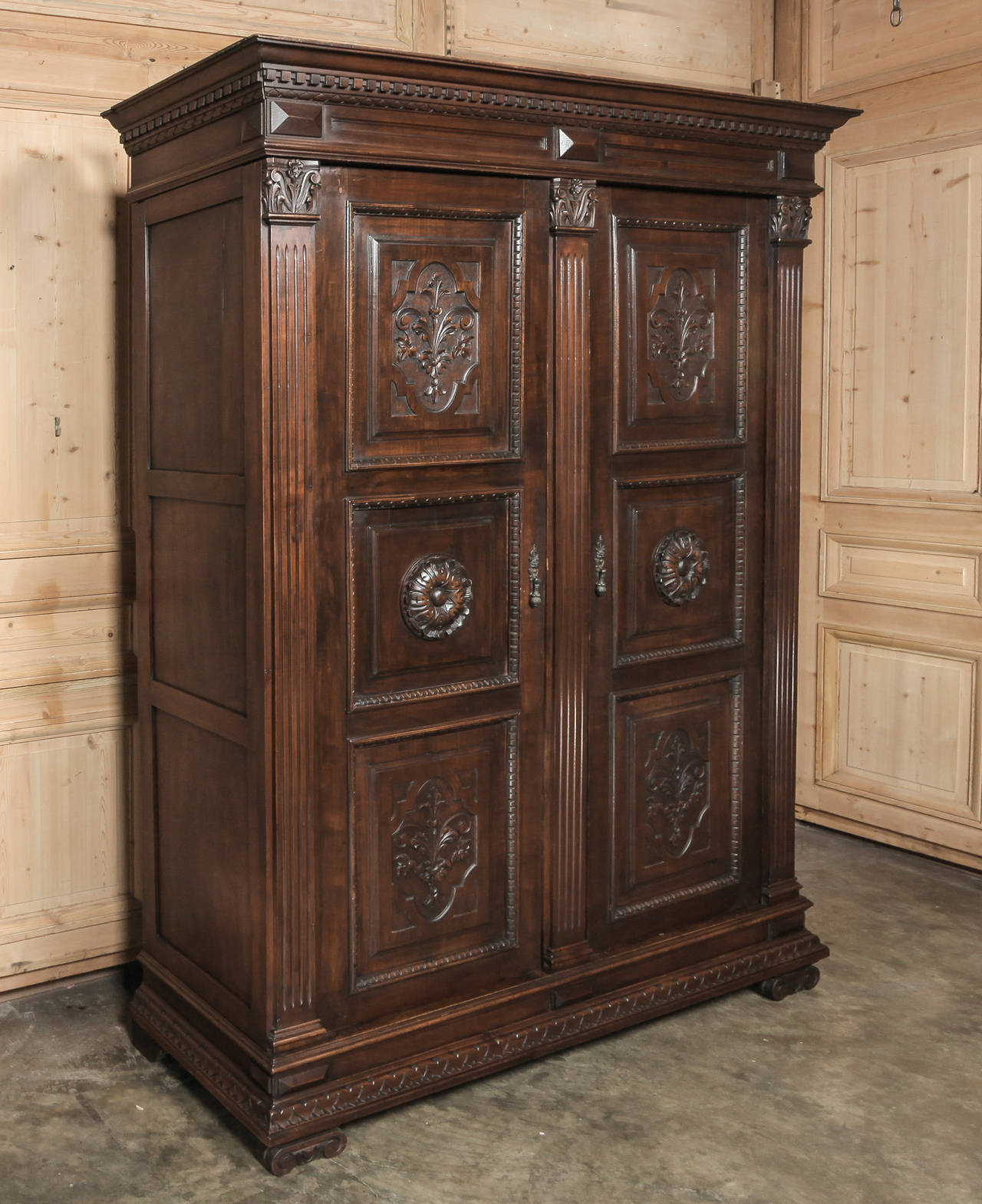Rendered in Renaissance magnificence from fine Italian walnut, this handsome armoire boasts fluted corner-posts topped with Corinthian corbels acting as framework for finely carved door panels arranged in threes. Bold rosettes at the center and