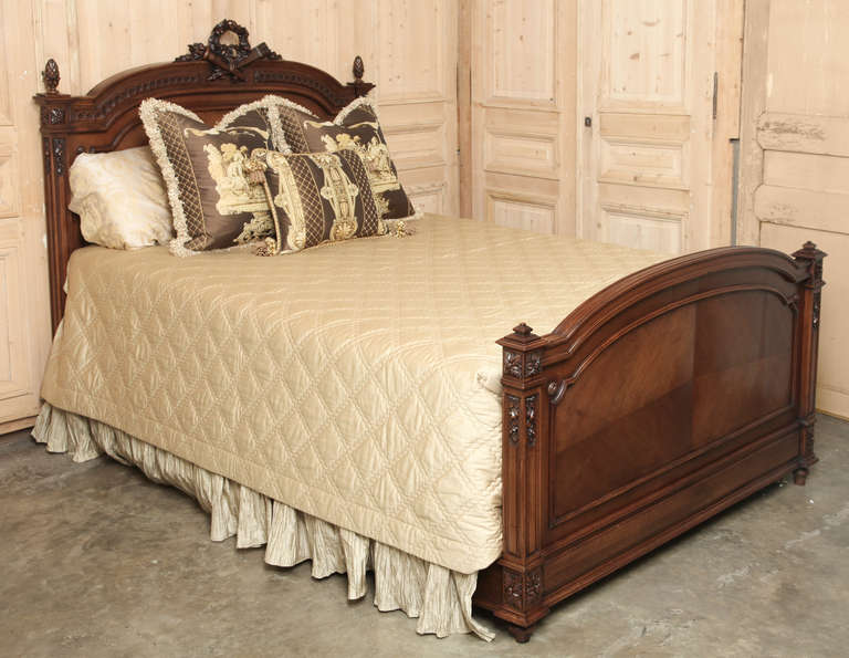 This Antique French Louis XVI Walnut Bed has been converted to accept a Queen Size Mattress Set!  Sculpted from fine walnut, it boasts classical architecture with cross-matched panels, fluted cornerposts, and a stunning floral wreath atop a torchere
