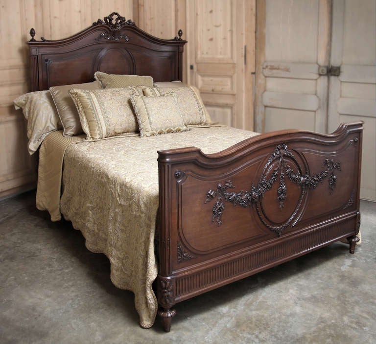 Hand-crafted from fine French walnut in the classic manner of Louis XVI, this elegant bed has been cleverly adapted to accept a Queen size mattress set! Featuring an elaborate ribbon motif sculpted atop the archer's bow crown, it features fine
