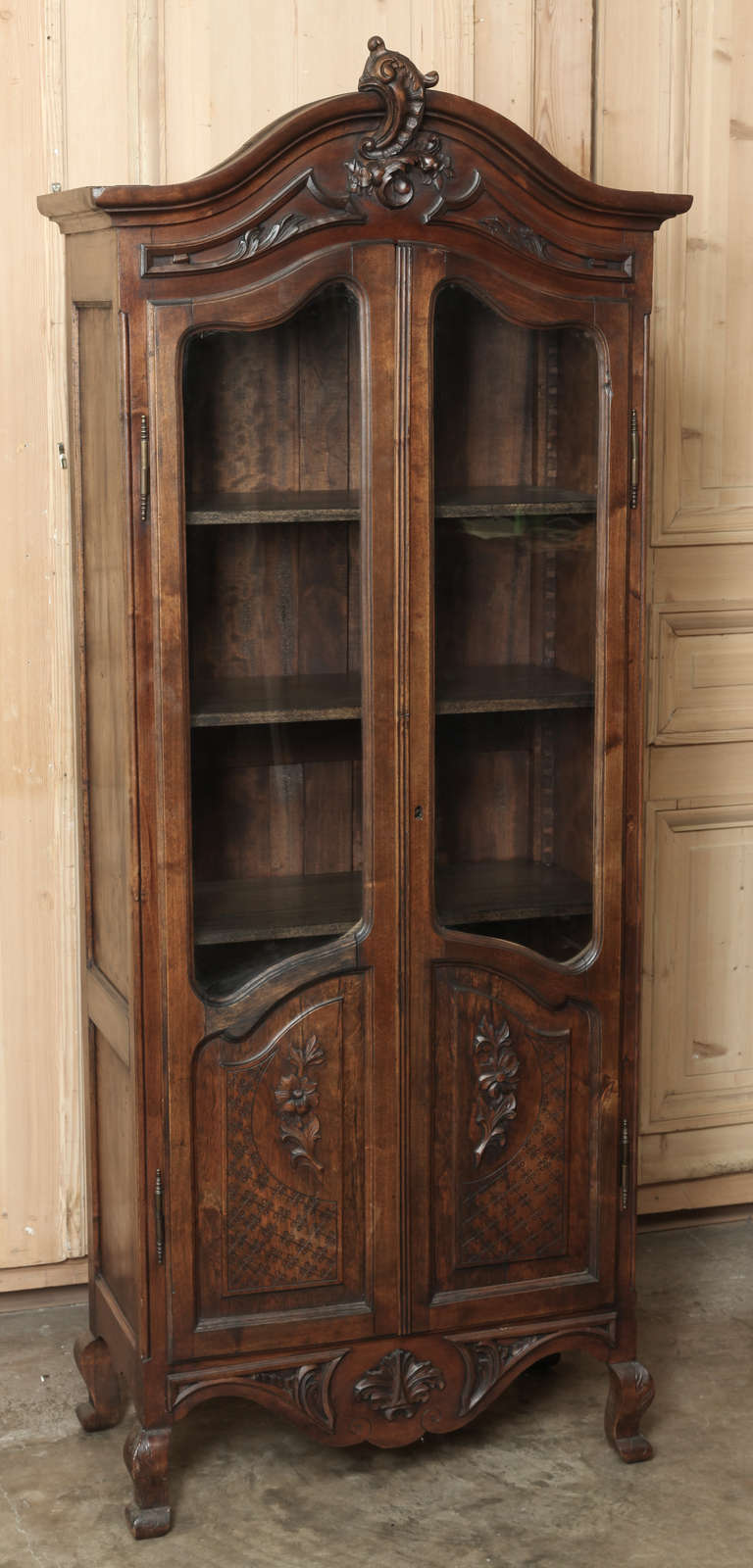 Featuring styling inspired by the rococo movement, a favorite of Louis XV, this Antique Country French Vitrine was hand-crafted from solid fruitwood, and features carved embellishments from the cartouche atop the arched crown to the boldly scrolled