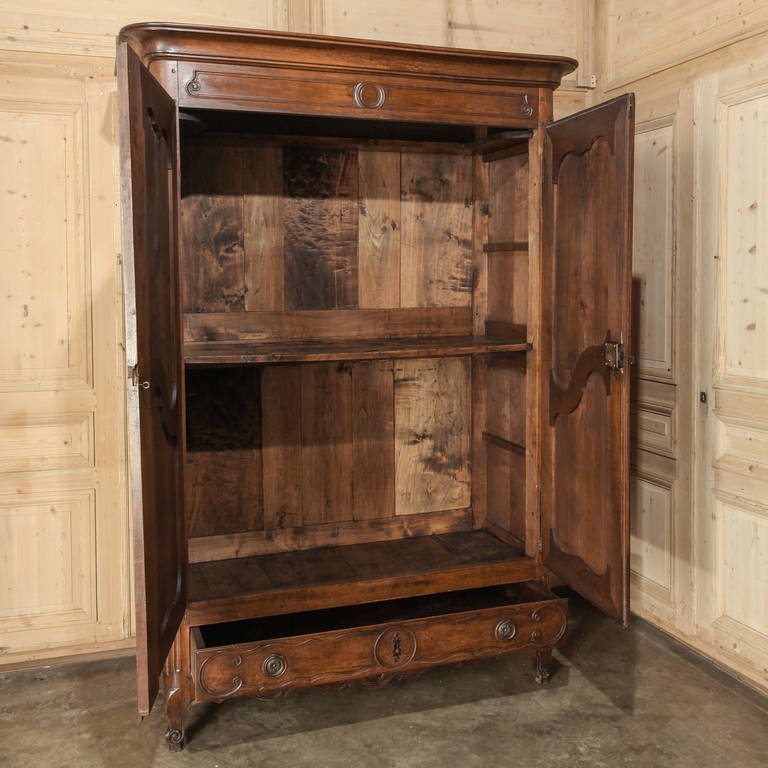 Rendered from fine French walnut by the master artisans of Provence, this handsome Country French armoire features a bold crown presiding over a subtly scrolled and molded framework, which includes a full width drawer below. Steel barrel hinges