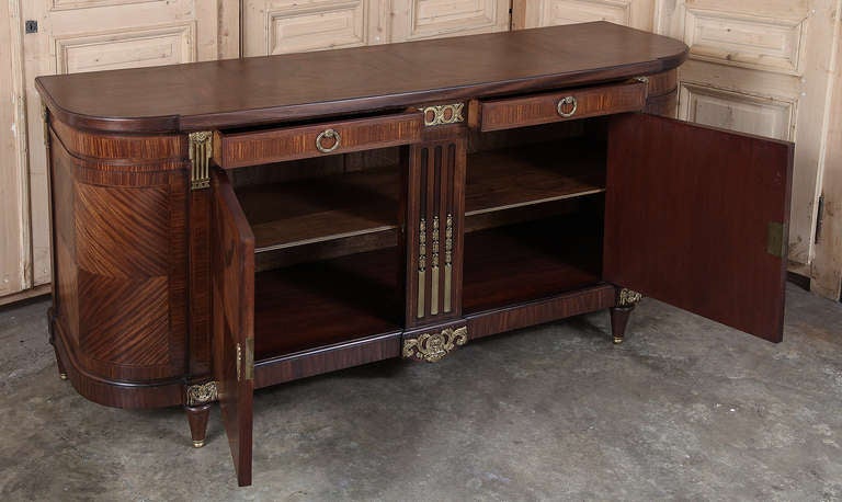 Lavished with exotic grain-oriented mahogany veneer, then inlaid with bordering and a stylized laurel garland, this buffet exhibits tailored architecture melded with opulent bronze ormolu mounts for an elegant effect. Rounded sides provide visual