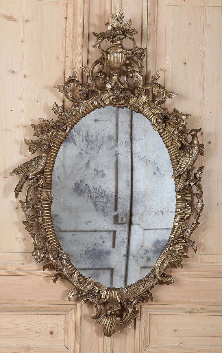Boasting an exuberant flair, this oval mirror features a hand-carved framework that is a masterpiece of sculpture, all finished in gold for an opulent effect. Fanciful urn, florals, foliates, even birds are depicted around the original mirror,