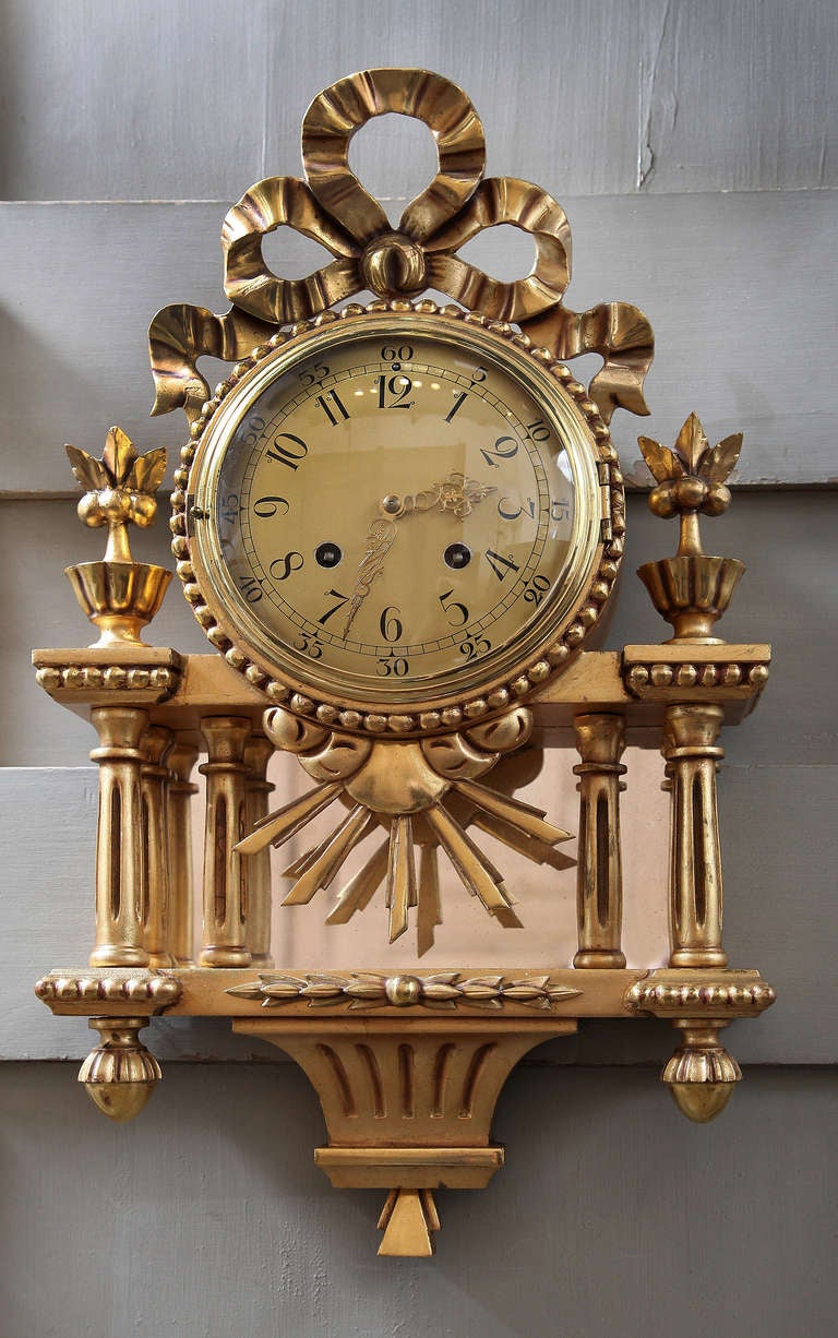 This regal looking clock was hand-carved and gilded to perfection, its finish to resemble bronze d'ore.  The combination of motifs is intriguing, with architectural detail, a flowing ribbon bow, columns, beading, and an interesting sunburst behind