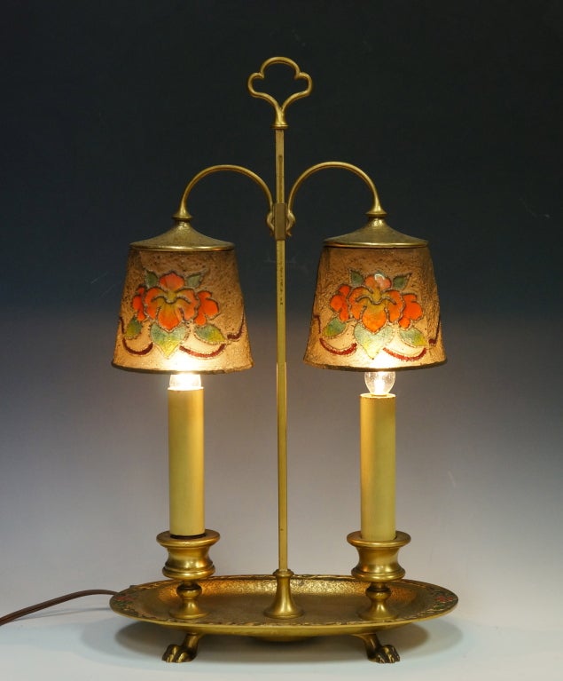This decorative Tiffany table or desk lamp features double candlestick bulb fixtures mounted with decorated mesh shades. It has a gilt bronze finish and base has enamel decoration. It is marked 