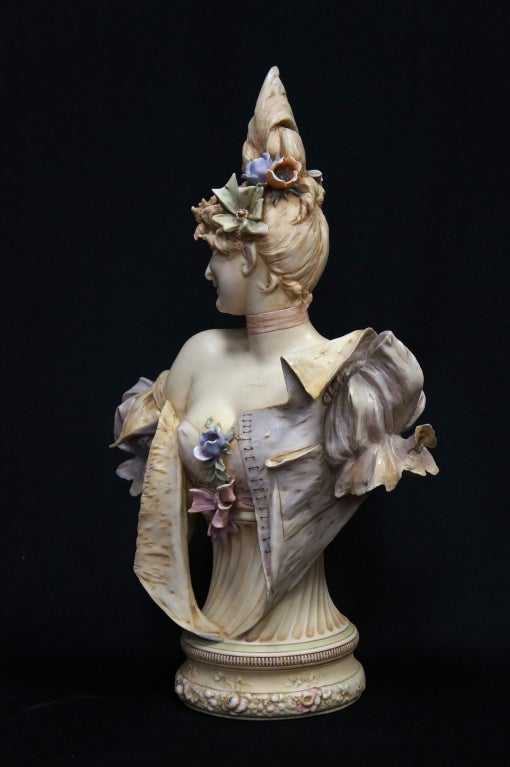 Beautiful art nouveau porcelain bust of a smiling young woman dressed in finery and wearing flowers in her hair mounted with a floral decorated base. It was manufactured by Reissner, Stellmacher & Kessel Amphora, a company known for very high