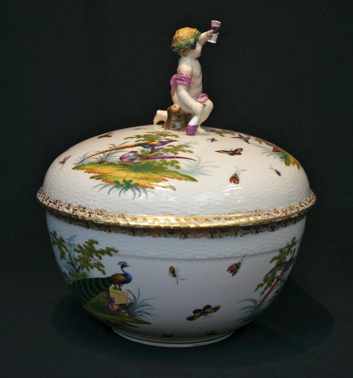The woven textured porcelain covered tureen is decorated with polychrome vignettes of animal life and flying insects. Borders are accented with gilt banding on bowl and lid. The lid is crowned with a figural finial of Bacchus lifting a wine goblet