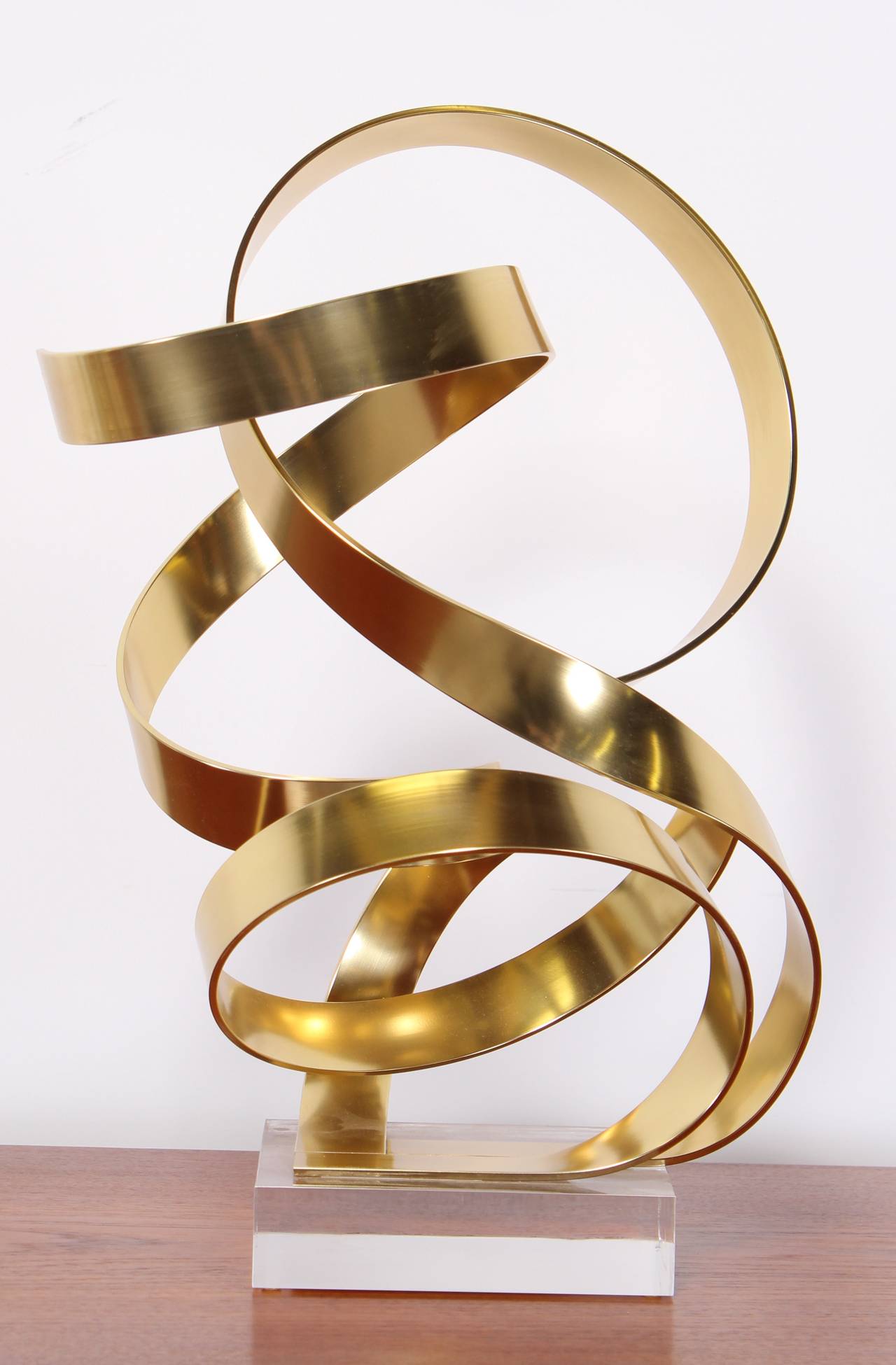 A whimsical gold tone anodized aluminum ribbon sculpture on a Lucite base signed by Dan Murphy 1985.