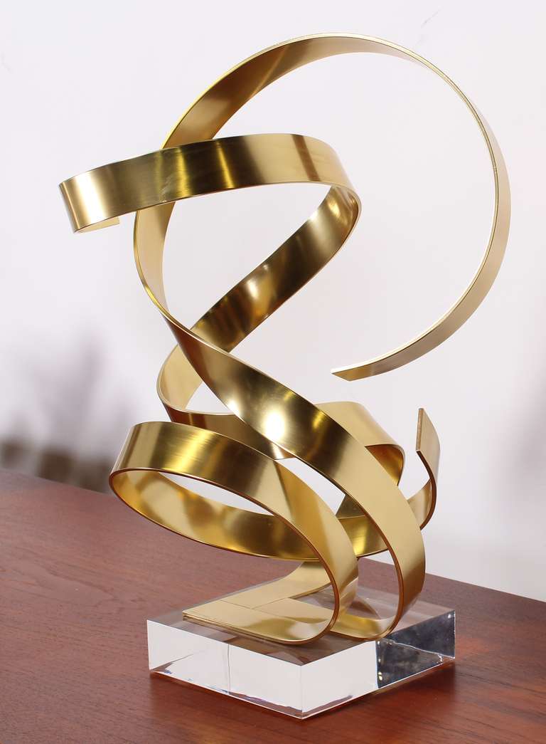 A whimsical gold tone anodized aluminum ribbon sculpture on a Lucite base signed by Dan Murphy 1985.