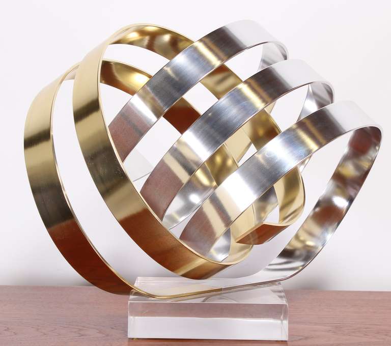 A whimsical gold and silver tone anodized aluminum ribbon sculpture on a Lucite base signed by Dan Murphy 1983.
