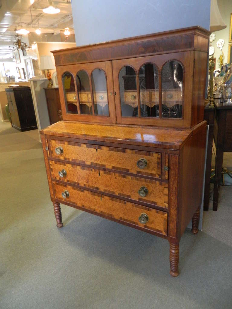 Large American Federal Period mahogany, curly birch and exotic veneer desk. Glass upper cabinets enclosing four small cabinets and niches over three lower pull out drawers raised on four spindle legs.
