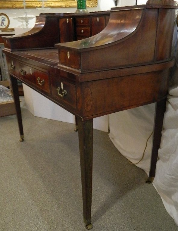 Mahogany cross banded Carlton House desk with satinwood and burl inlay and bone escutcheons. It has a pull out leather top writing surface that tilts to support a book or sheet music.  The brass lock hardware is signed by the English maker and the