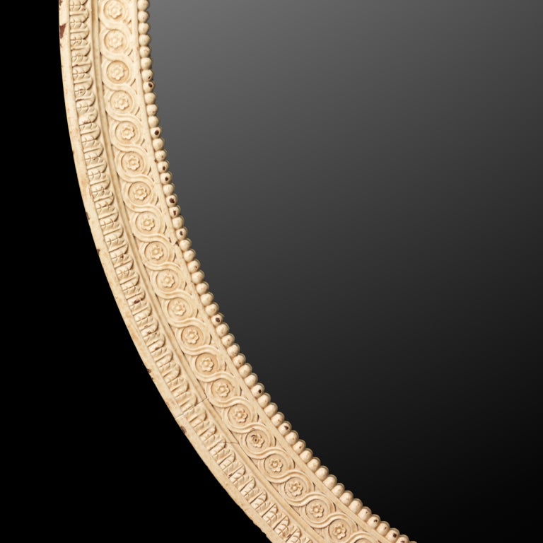 A pair of carved wood oval mirrors in the style of George III and the late eighteenth century. The moulded frames are enriched with carved guilloche decoration, flanked by an inner bead moulding and an outer waterleaf, all within a square moulded