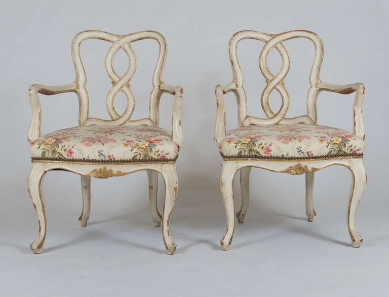 Very Elsie de Wolfe chic.  Based on English Georgian designs.  Each with interlaced back issuing shepherd's hook armrests; on cabriole legs.  With gilt decoration and detailing.  Note extensive losses to paint.