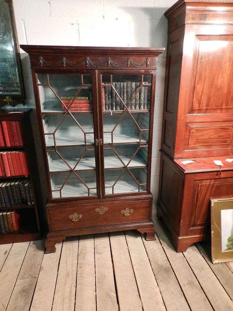 Small mahogany bookcase with 2 astragal glazed doors.  The frieze has carved swag decoration.  The upper bookcase section sits upon the base which has one drawer with brass handles and is supported by shaped bracket feet.  The interior has 3