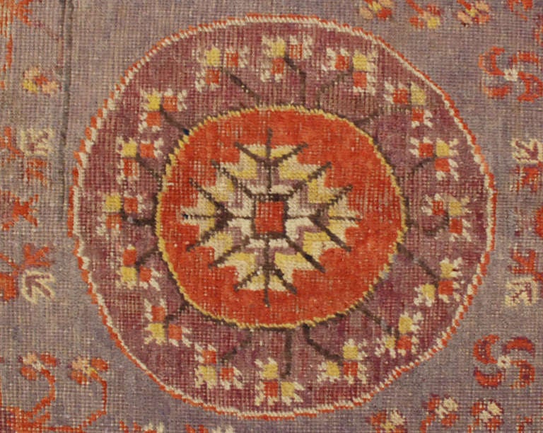 Un antique tapis Khotan d'Asie centrale avec un médaillon central asymétrique au milieu d'un champ de fleurs sur un fond violet, entouré de multiples bordures complémentaires.

4'1