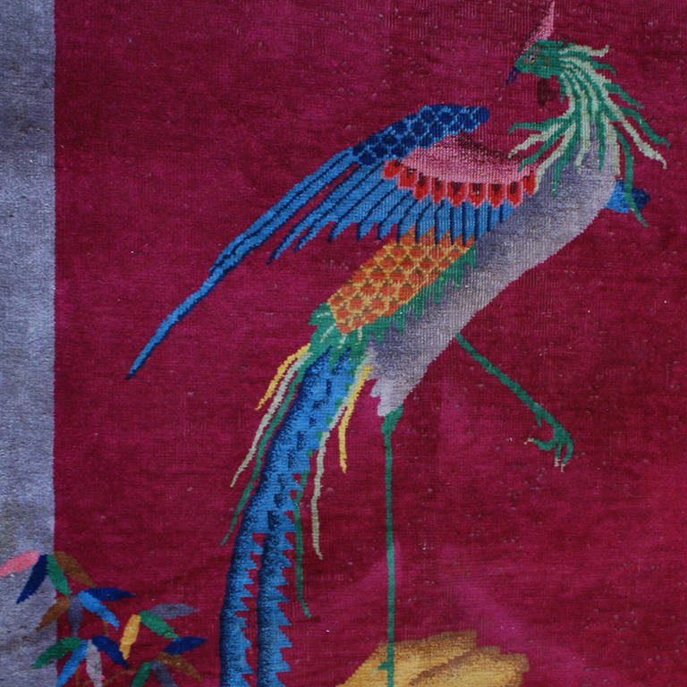 An early 20th century Chinese Art Deco rug with asymmetrical pattern of a phoenix amidst a garden landscape on a cranberry background, surrounded by an indigo border.
