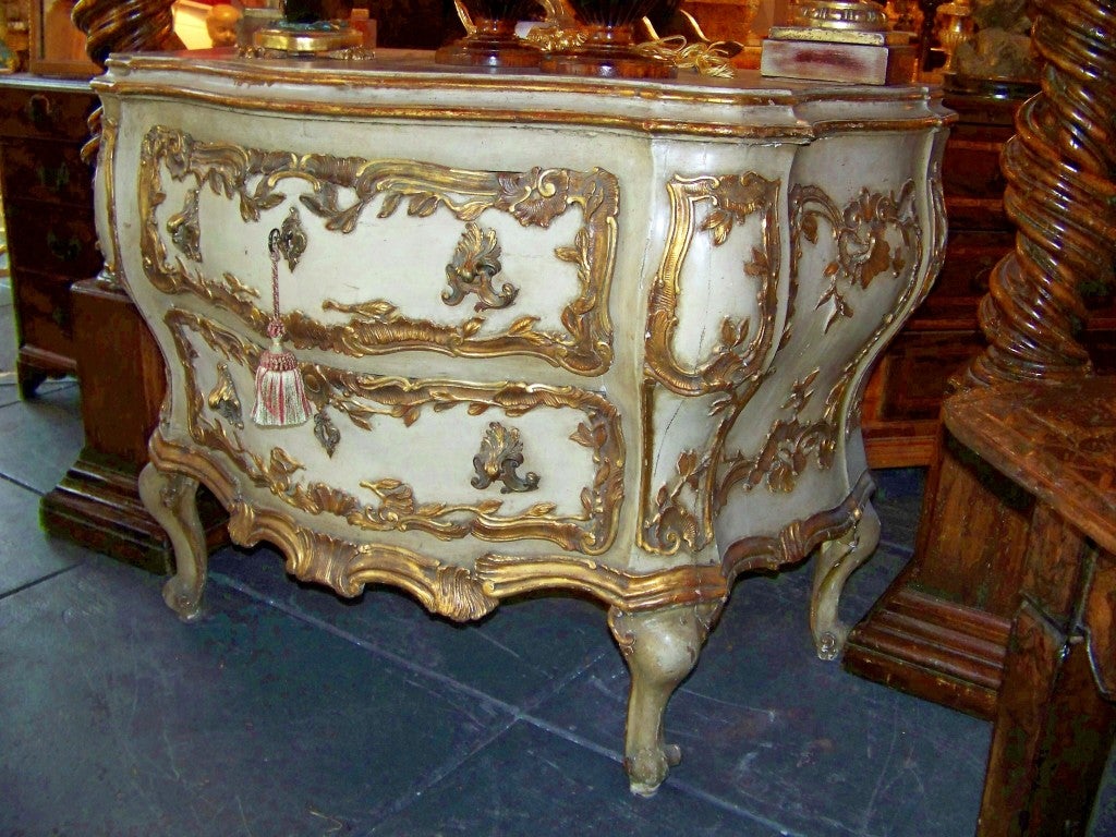 An unusual creme painted and giltwood decorated Venetian bombe' commode in the 18th century rococo style . This chest with unusually pronounced curves and large gilt bronze shell form pulls . The top a nicely worn faux with a gold wash