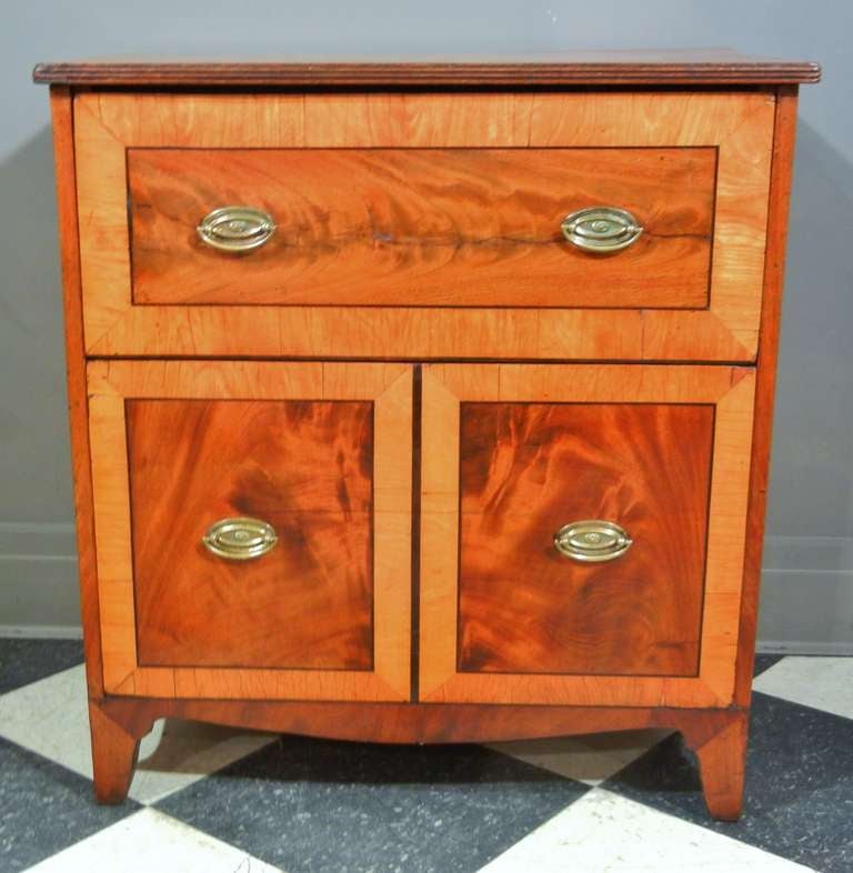 This versatile little chest has one drawer over a deeper drawer. The bottom drawer is veneered to look like cupboard doors, which gives the chest a clever twist. The figured mahogany veneer and the satinwood banding provide a luminous, cheerful
