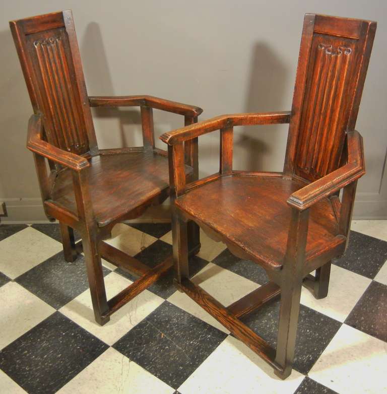This pair of French, studio-made Renaissance-Revival arm chairs have hand-carved linenfold panels set in tall narrow backs, and have trapezoidal seats, U-shaped arms, cupid's bow skirts, and beveled, cantilevered legs on H-shaped stretchers with