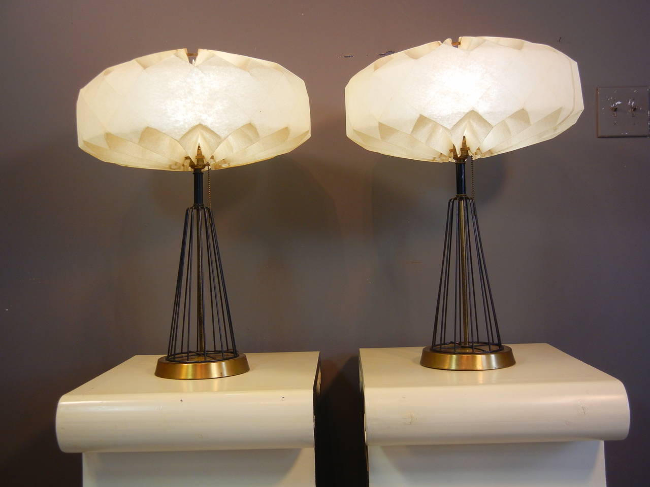 This pair of futuristic table lamps have round brass basses, shafts of black iron rods in an upside-down cone configuration, and Fiberglass flying saucer-shaped shades.

The shades are pleated top and bottom where they are attached to brass rings,