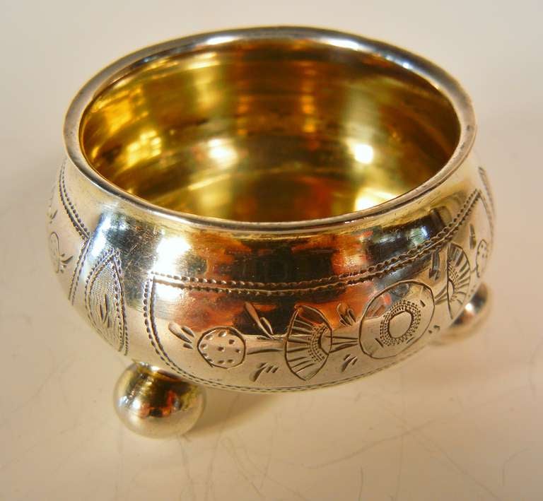 The salt cellar is engraved with a traditional Russian stylized floral motif around the exterior of the bowl, it sits on three ball feet, and the interior is gilded. The hallmarks are 