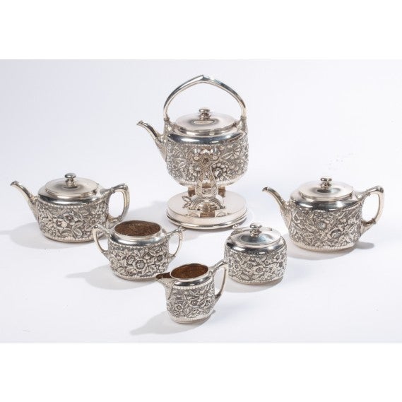 Seven pieces including hot water kettle with hinged lid, kettle stand with burner and hinged lid, two teapots with hinged lids, open creamer, sugar bowl with detachable lid, open waste bowl, all with deep repousse work and chasing. Signed on the