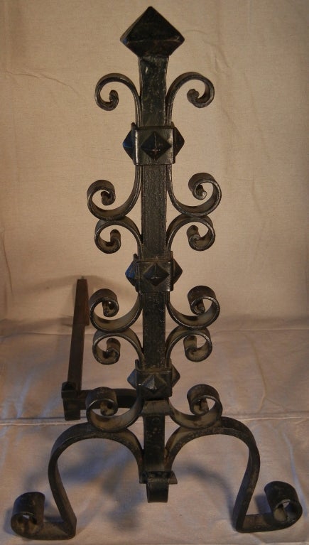 This pair of Arts & Crafts period andirons are handmade of wrought and hammered iron, and feature diamond tops, with scrolls, diamonds, straps and loops along the square shafts, on half-heart shaped legs with scrolled feet. They were made for a