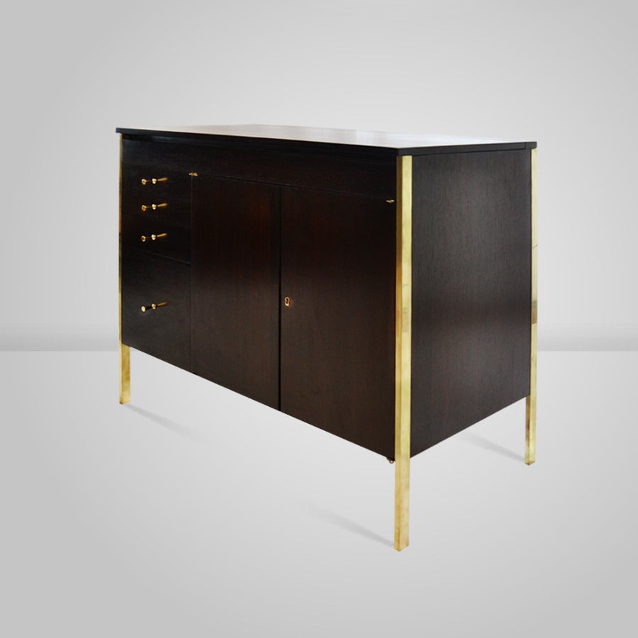 Handsome and compact bar cabinet designed by Paul McCobb for his Connoisseur Collection and produced by H. Sacks and Sons. The top lifts up to create a working space and storage. Refinished in a beautifully rich dark brown. Brass legs and signature