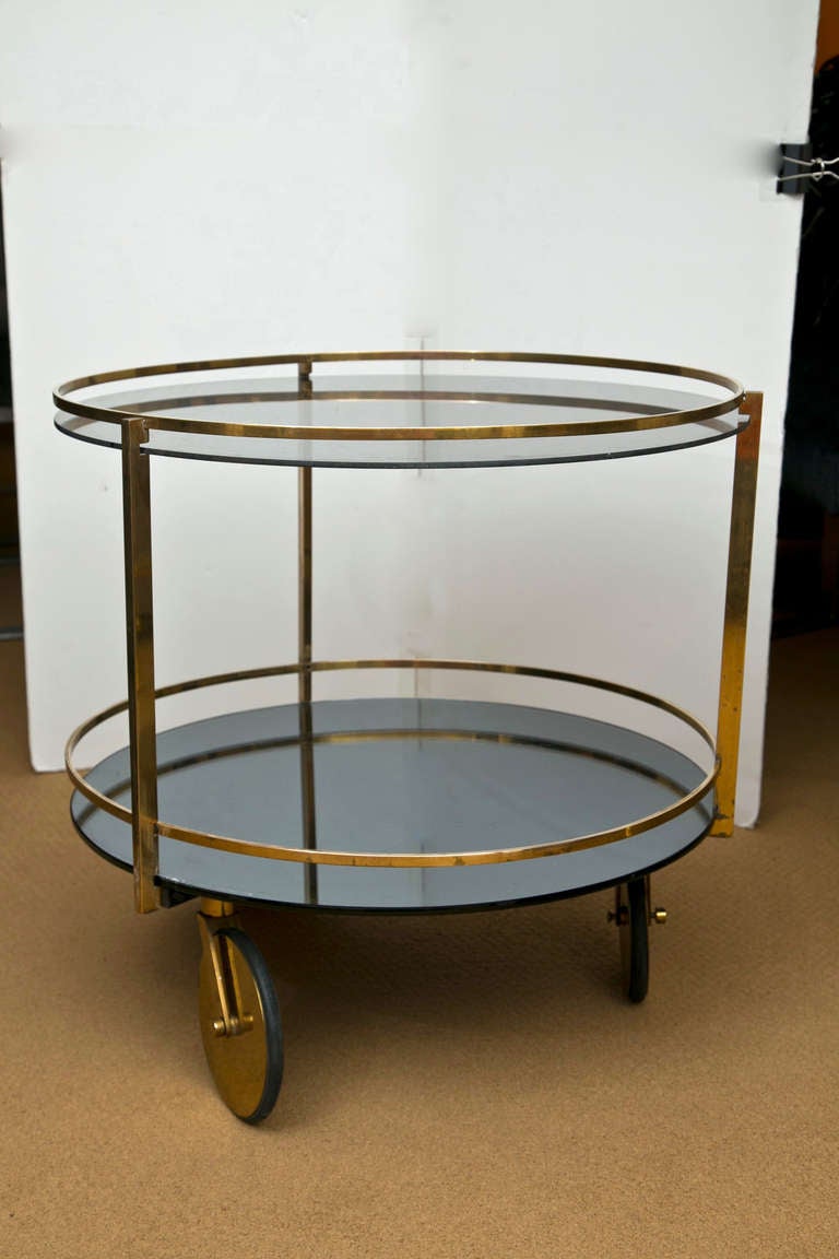 Iconic and rare solid brass double level cart on oversized wheels. The top level is smoked glass and the lower is smoked mirror. Both the glass and mirror have age appropriate scratches expected to be found in a piece of this vintage.