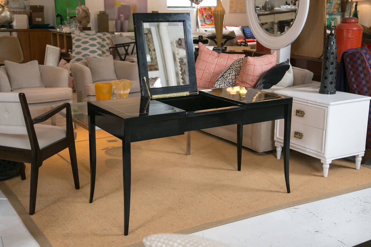 Gracefully designed vanity in the style of Tommi Parzinger. Finished with ebonized lacquer and delicate gold-leaf detailing. Constructed with 2 drawers, and a hidden compartment atop which opens to reveal a mounted mirror. 

Extra 23