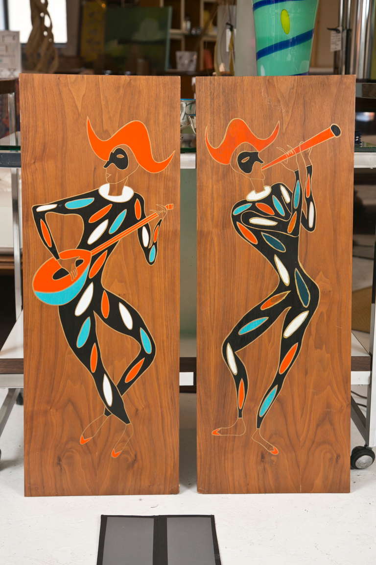 A pair of mid-1950s painted wood panels depicting instrument playing masked minstrels in colorful Harlequin style dress. One plays a horn, the other a guitar or mandolin. Old reworked corners, minor scuffing.