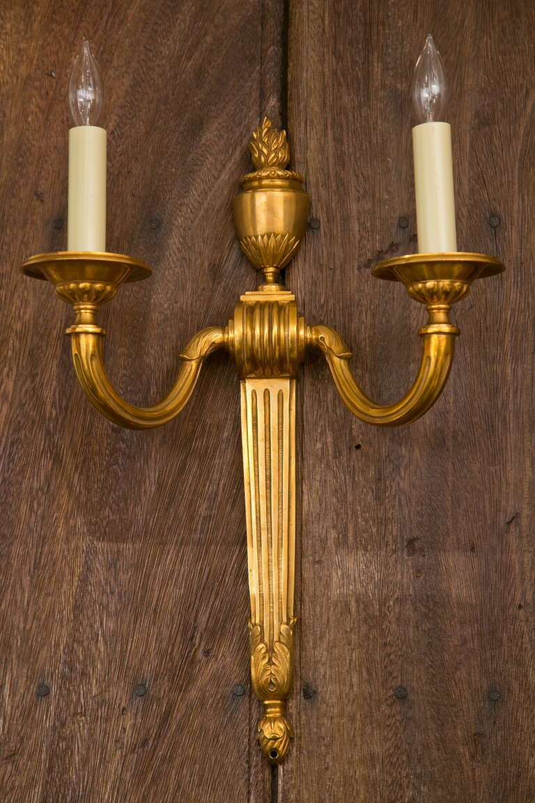 Very much in the manner of Caldwell Lighting, this gilt bronze sconce pair has over-all fluting, generous proportion and weight, and a flame urn finial.
