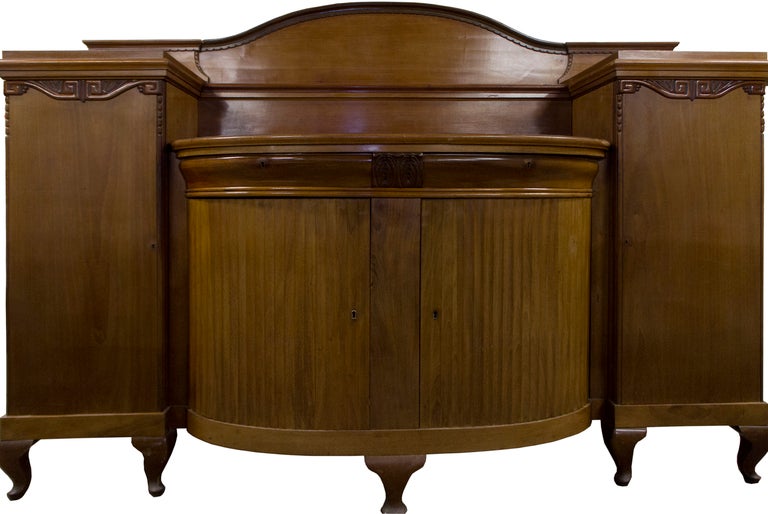 Designed to be the centerpiece of an early 20th century dining room and crafted of solid blonde mahogany and fir, the buffet features several elevated serving areas, including a pair of curved, locking drawers, a long upper shelf, and a myriad of