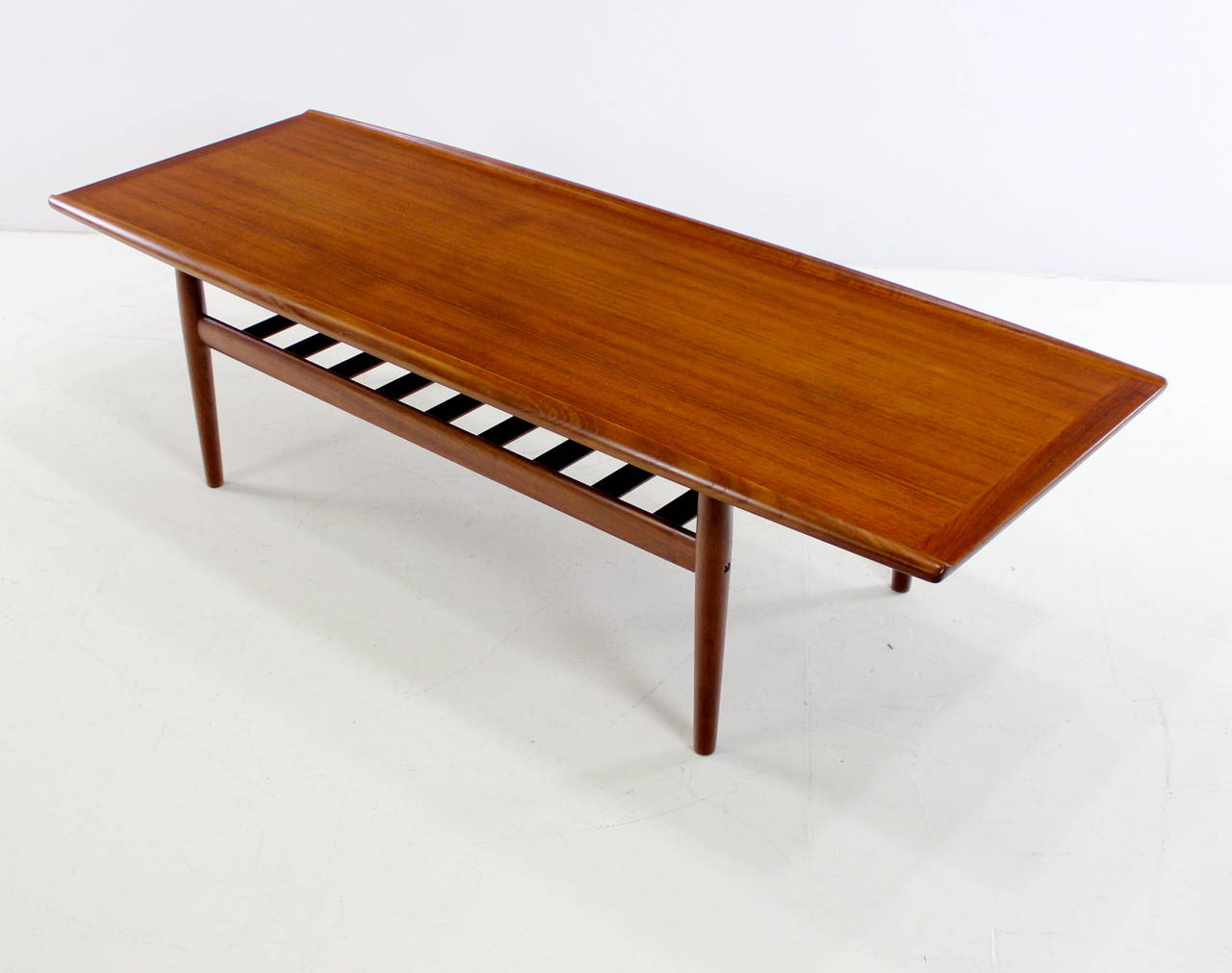 Danish modern coffee table designed by Grete Jalk.
Solid teak.
Superior Jalk style and quality with inlaid, upturned table edges and lower slatted magazine shelf.
Professionally restored and refinished by LookModern.
Matchless quality and