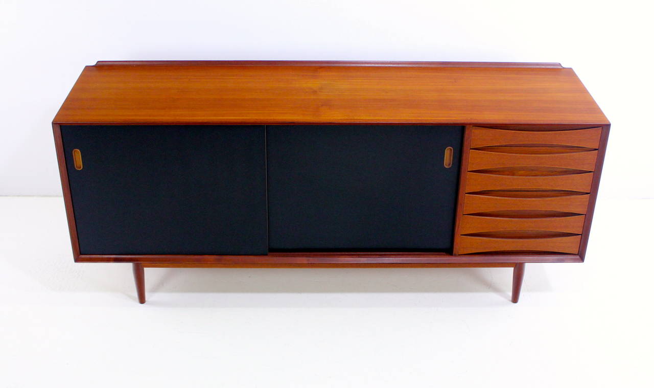 Danish modern credenza designed by Arne Vodder.
Sibast, maker.
Black sliding doors open to adjustable shelving in center and left.
Six generous drawers on right.
Semi-finished on the back for possible 360 degree use.
Richly grained teak with
