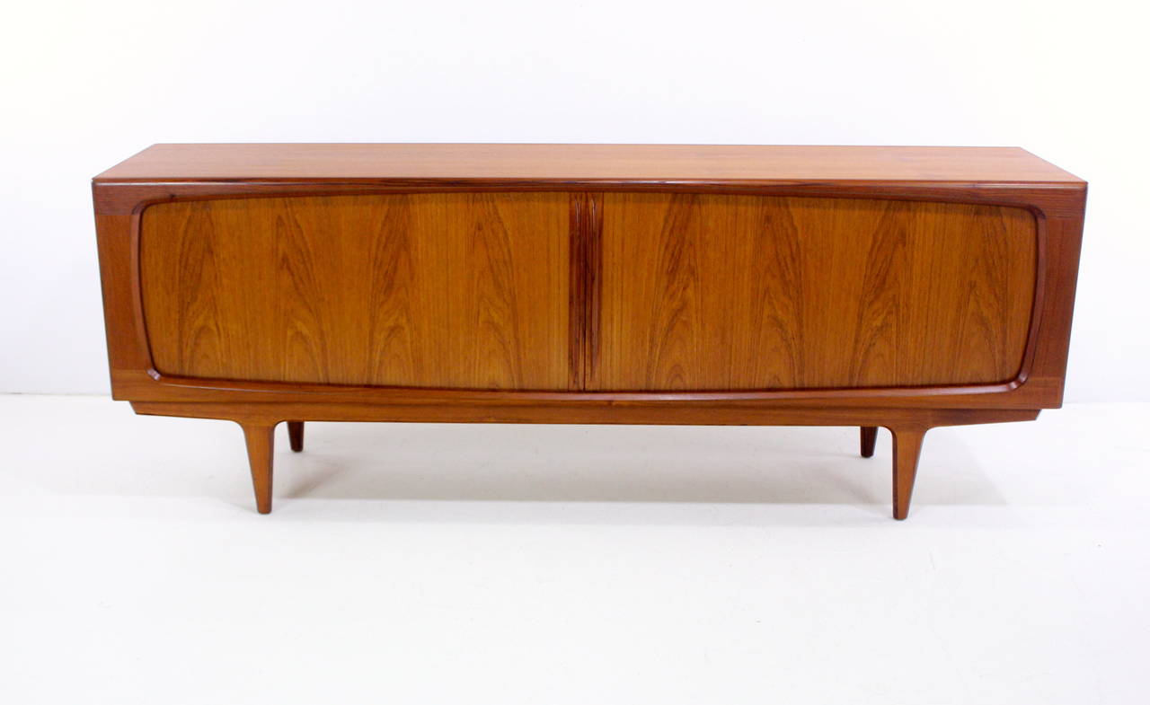 Danish modern credenza designed by Bernhard Pedersen & Son.
Richly grained teak.
Voluptuously curved frame surrounds tambour doors, which glide open to adjustable shelves on each end. 
Three drawers in the middle. 
Perfectly styled, scaled base