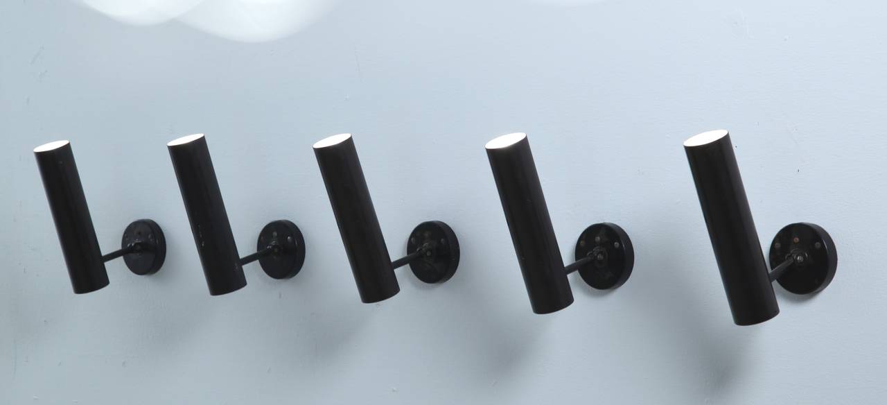 A set of 5 slim, long cylindrical black metal ceiling lamps. The spots are attached with a black ceiling mount and are adjustable.