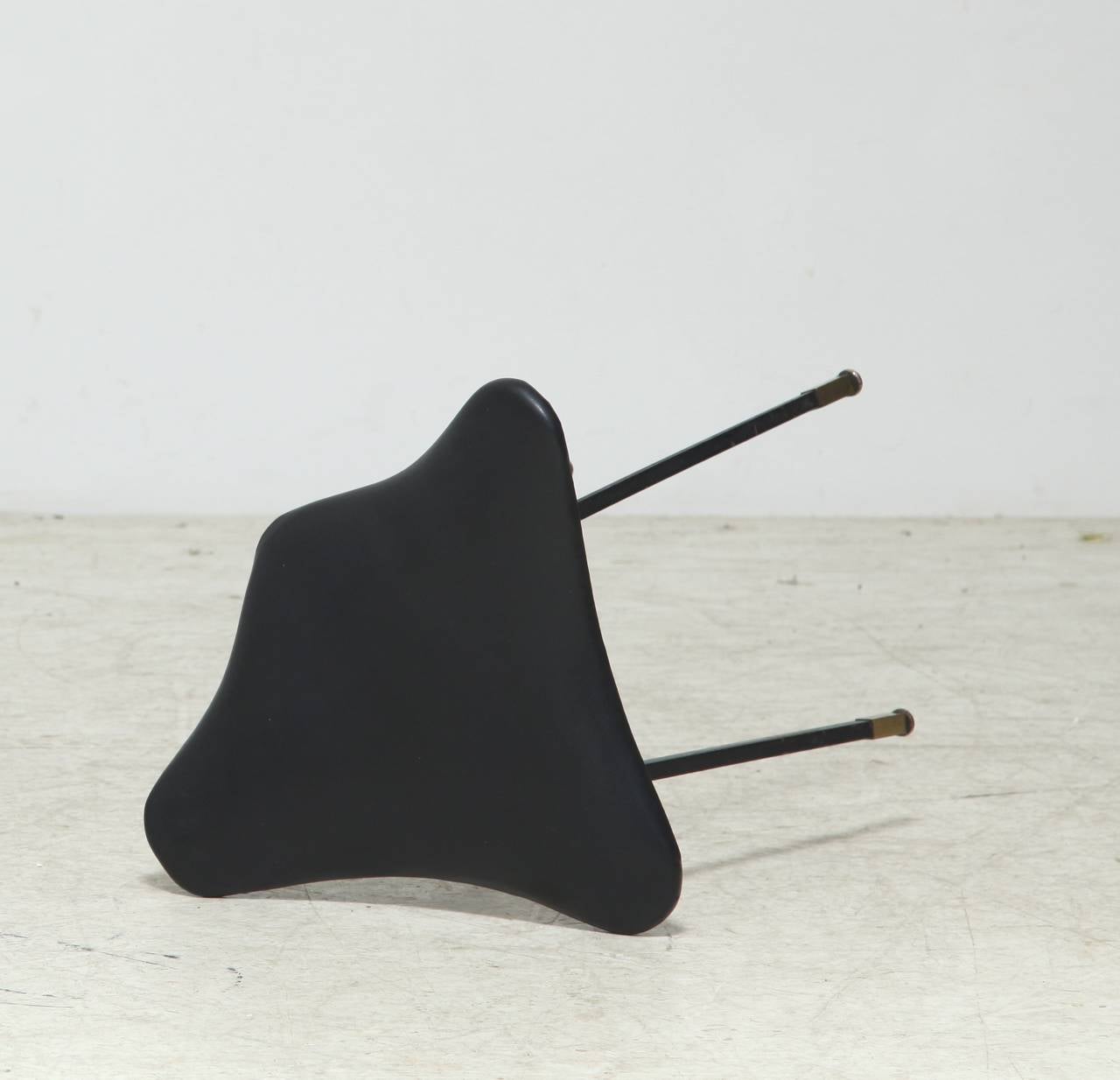 A small Jacques Quinet tripod stool, made of metal legs with brass feet and a saddle-shaped seating made of slightly bent wood with a black leather upholstery.