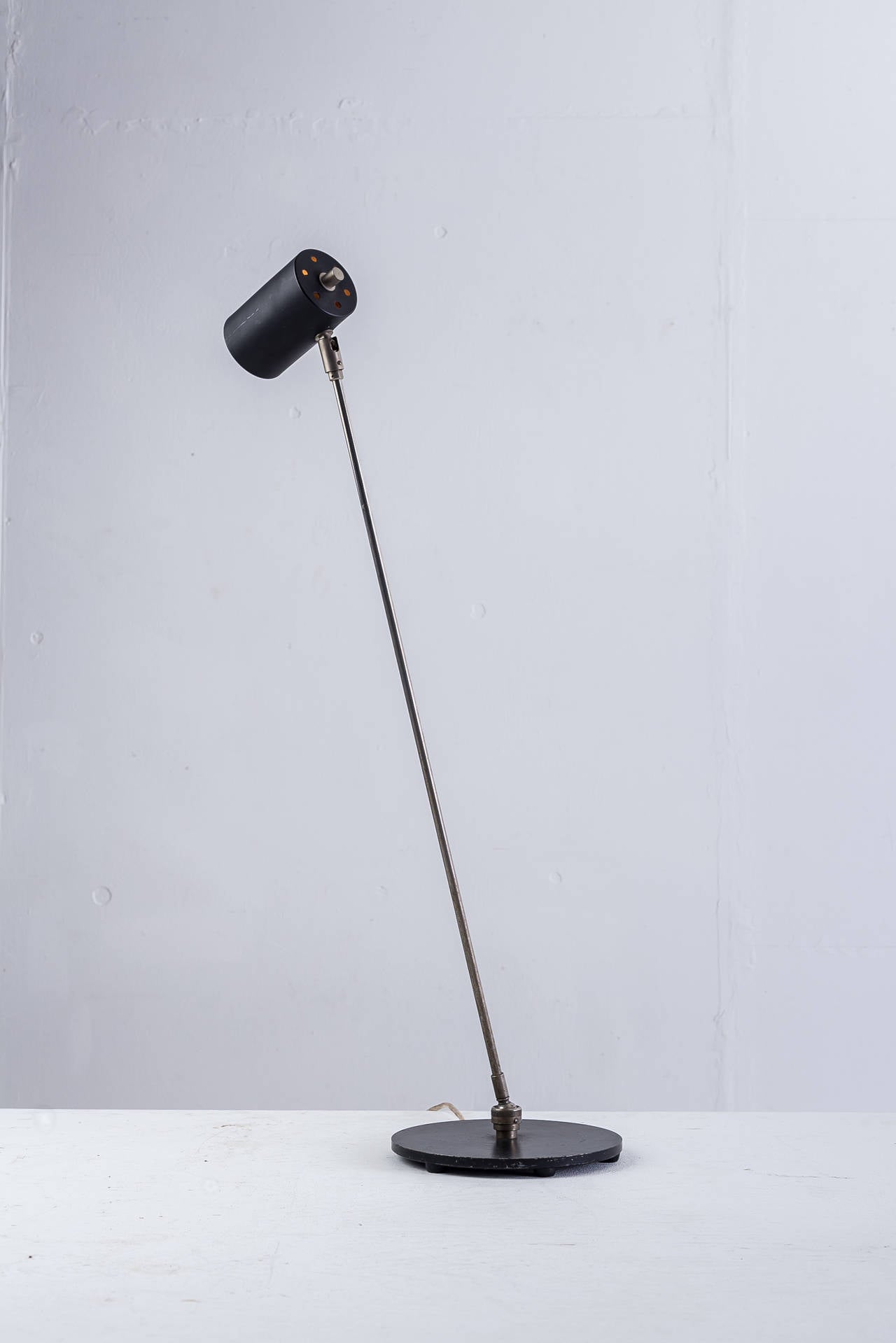 A modernist floor lamp from the 1950s, made of a black lacquered metal base and cylindrical shade on a brass stem. The ball-joint construction at the bottom of the stem makes the lamp adjustable at a 360 degree angle. The angle of the hood is also