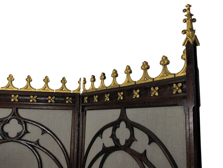 An English oak room divider in the Gothic manner comprising two large tracery panels with castellated top border and finials in gold leaf. The panels upholstered in grey linen can be folded in either direction.